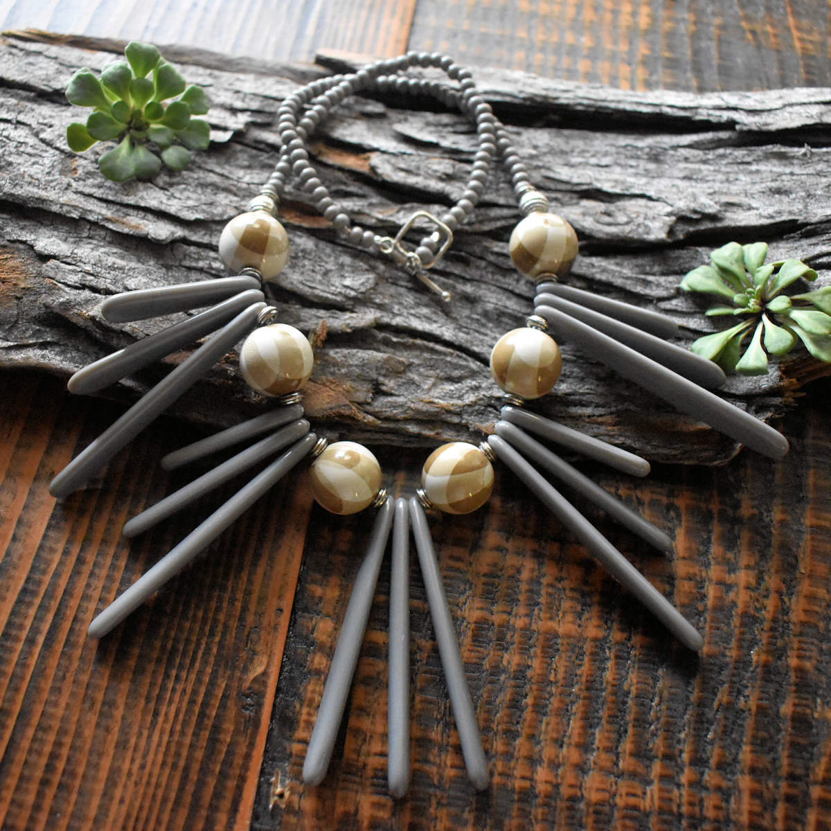 A necklace with beige rounds and gray long drop beads draped across a wood background. The necklace has five sections of long gray beads; the center section is three of the longest beads and the two sets on each side are graduated from long to short. Between each section are beige beads that have geometric arc design on them. 