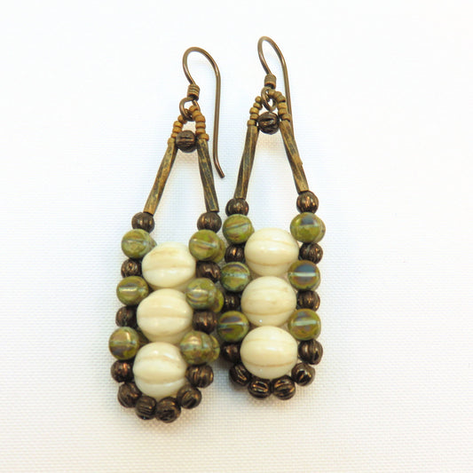 Cream and moss earrings with dark ear wires lay on a white background. These earrings each have three cream colored melon beads that are secured by an outline of mossy green and bronze beads. 