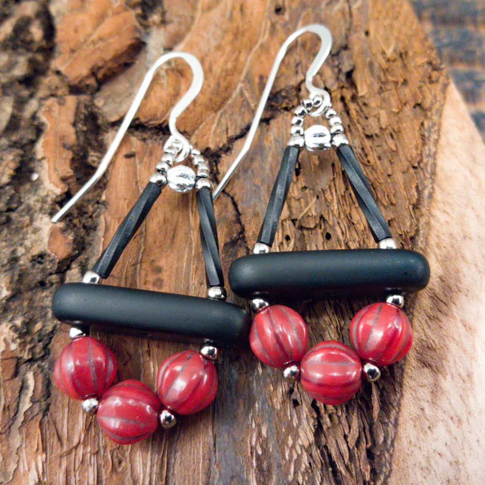 Red and black earrings lay on a piece of bark. The earrings have a black triangle formed from two upright black tubes and a matte bar at the base. Below the bar is an arc of three red ridged round beads.