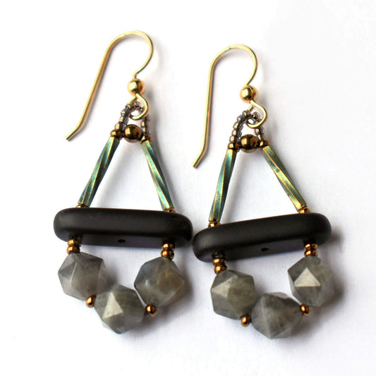 Black and gray earrings lay on a white ackrgound. The earrings have a  triangle formed from two upright iridescent green tubes and a matte bar at the base. Below the bar is an arc of three gray geometric cut stone beads. 