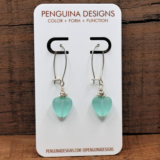 A pair of earrings on a white card rest against a wood background. The earrings have long silver oval wires that latch and small sea glass turquoise hearts at the bottom.