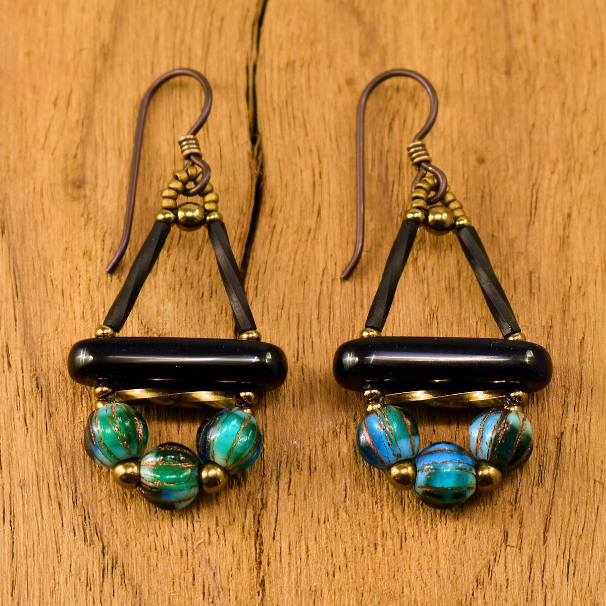 Black earrings with blue and green variegated beads and dark ear wires on a wood background. These earrings are shaped like a triangle with a rocker bottom.  The top of the rectangle is formed by twisted matte black tube beads and the bottom is a glossy black bar. Below the bar is a horizontal gold tube, and at the bottom is an arc of ridged round beads that have internal swirls of green, white, blue, and turquoise.