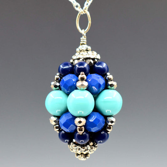 A big pendant hangs against a gray background. The pendant is formed from a center row of turquoise rounds, and then two increasingly dark blue rows. A vertical row of silver beads extends from the turquoise beds to the top and bottom. The pendant is capped with a silver finding. 