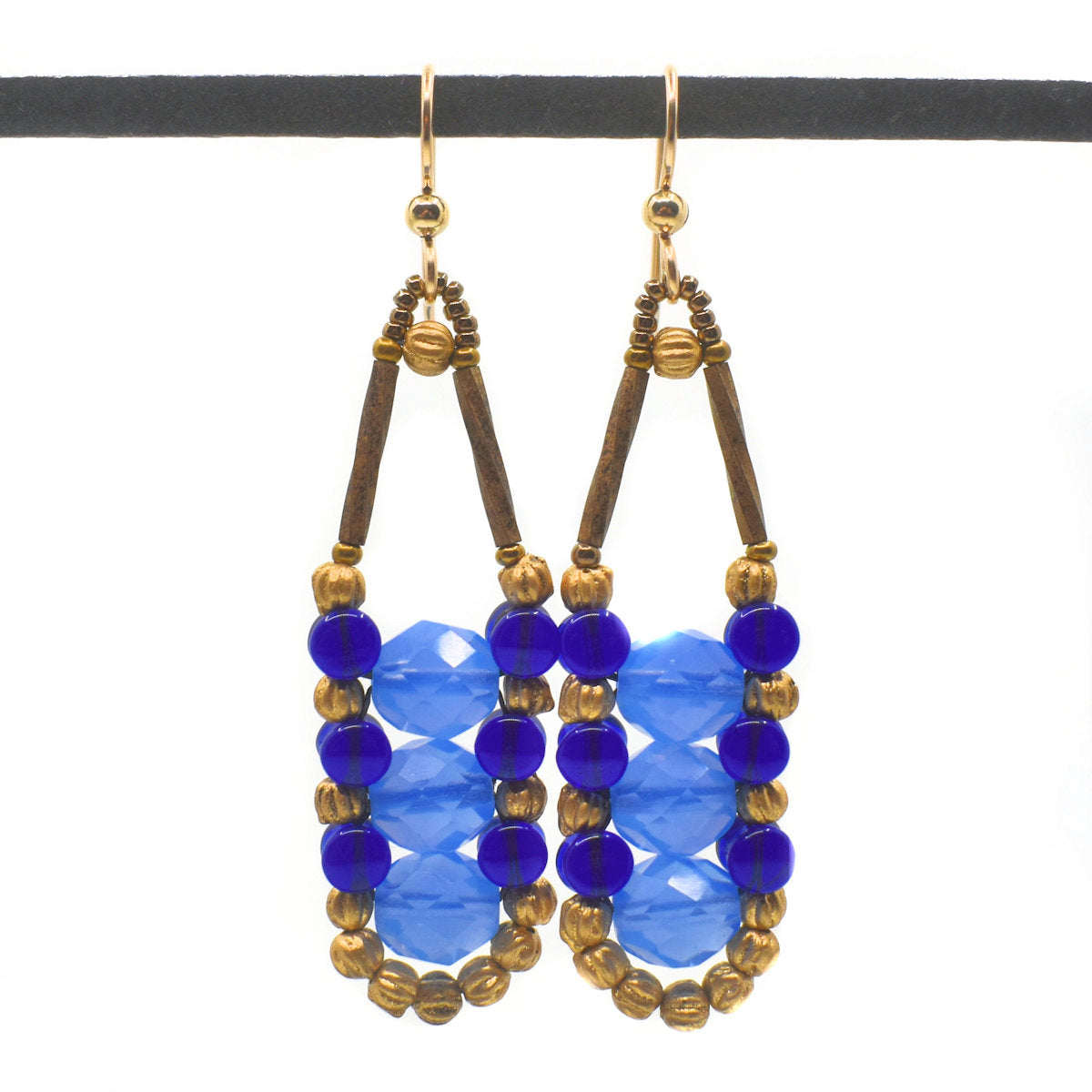 Translucent light and dark blue earrings with gold accents and wires hang from a black bar against a white background. These earrings each have three semi-transparent light blue rounds held in place by an outline of transparent cobalt and gold beads.