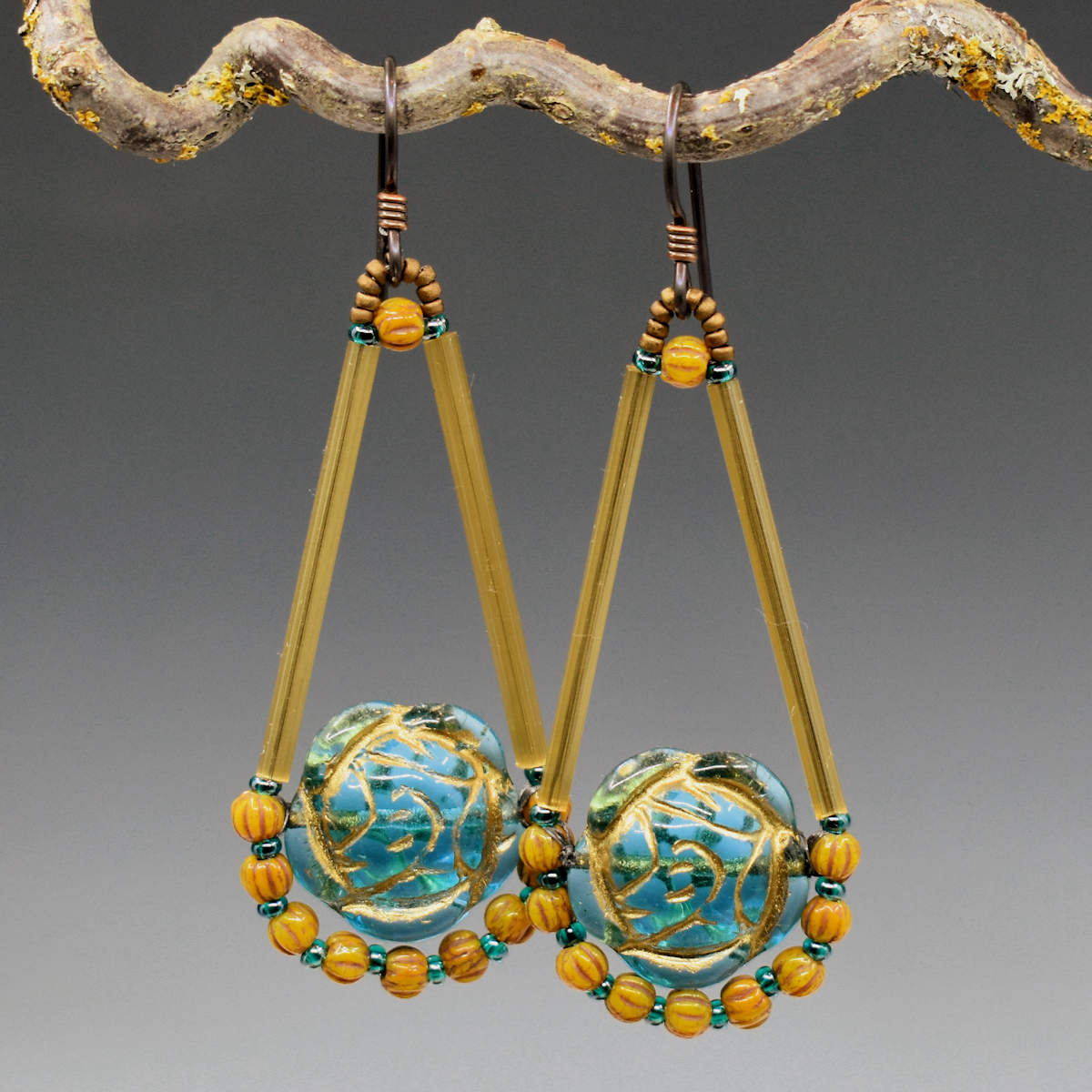 Long gold and aqua teardrop shaped earrings with dark ear wires hang from a lichen covered twig. The teardrop's widest part holds a transparent aqua rose beads with gold details.  The outline of the teardrop shape is formed from gold satin glass tubes with a swag of dandelion melon beads forming the bottom curve.. 