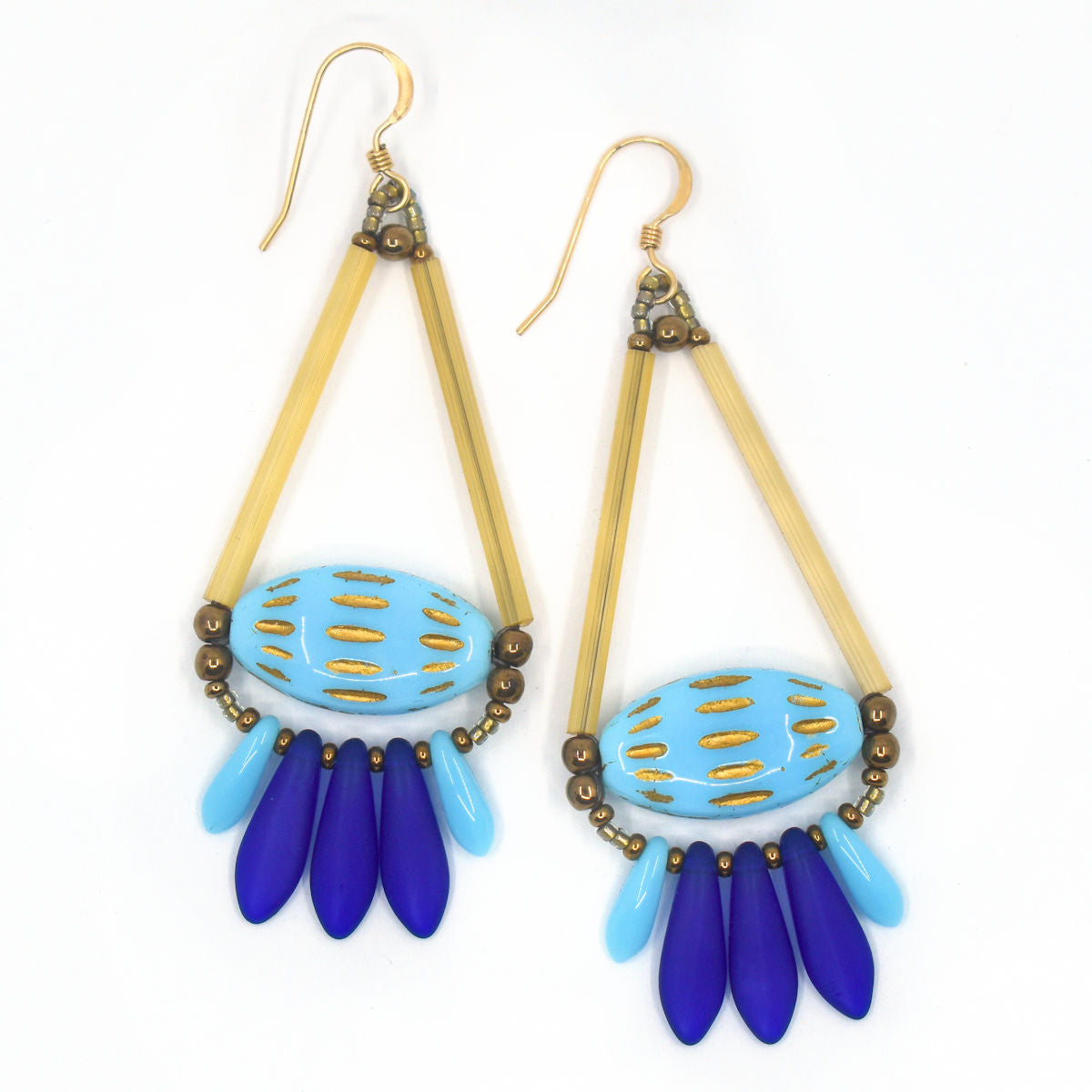 Wide blue earrings lay on a white background. The earrings are a wide teardrop shape with a fringe of matte royal blue and glossy sky blue daggers at the bottom. Across the middle of the earring are elongated light blue ovals with rows of gold embossed dashed lines across them. The triangle sides of the earring’s teardrop shape is formed by long gold tube beads 