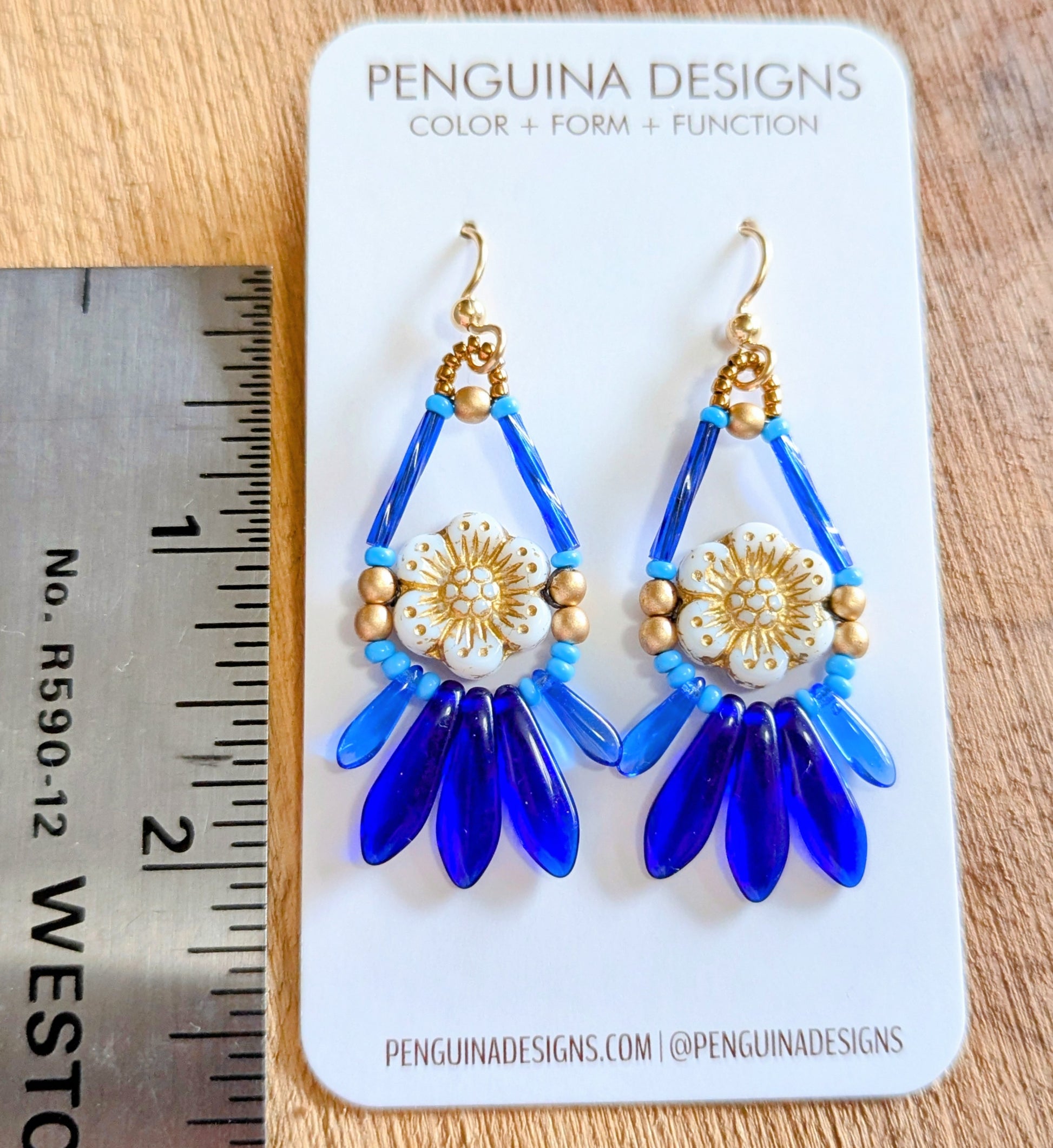 Beaded earrings with a white flower surrounded by blue and gold beads on a Penguina Designs card with a ruler for scale.