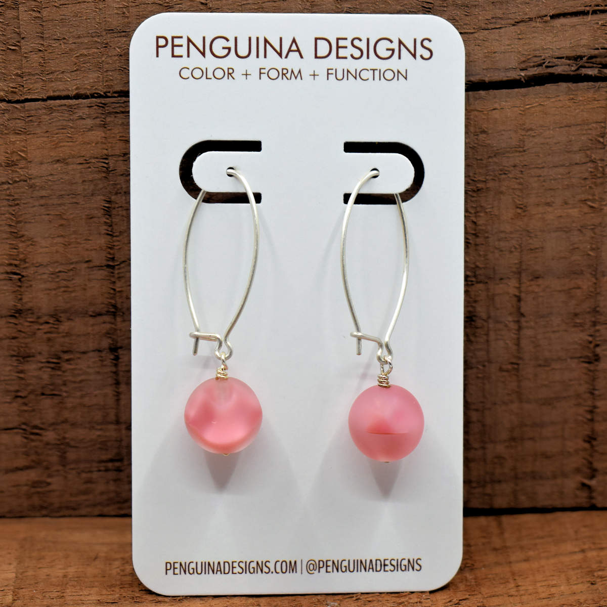 A pair of earrings on a white card rest against a wood background. The earrings have long silver oval wires that latch and frosted pink glass balls at the bottom.