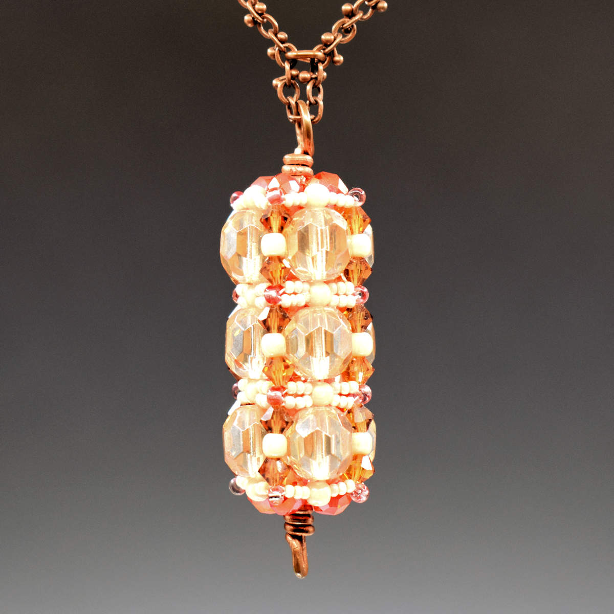 A peachy colored pendant hangs from copper chain. The pendant has three rows of pale yellowy peach with horizontal rows of cream seed beads between them. There is a vertical row of orange crystal beads that runs between the largest peachy yellow beads. 