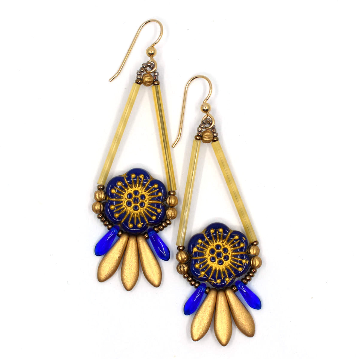 Gold and dark blue earrings with gold wires lay on a white background. The earrings are teardrop shapes, with the top formed by long gold tube beads. The circular part of the drop is filled with a dark navy blue flower with embossed gold details. Below the teardrop is a fringe of dagger beads, small blue ones on the outer edges and three larger matte gold daggers in the middle.