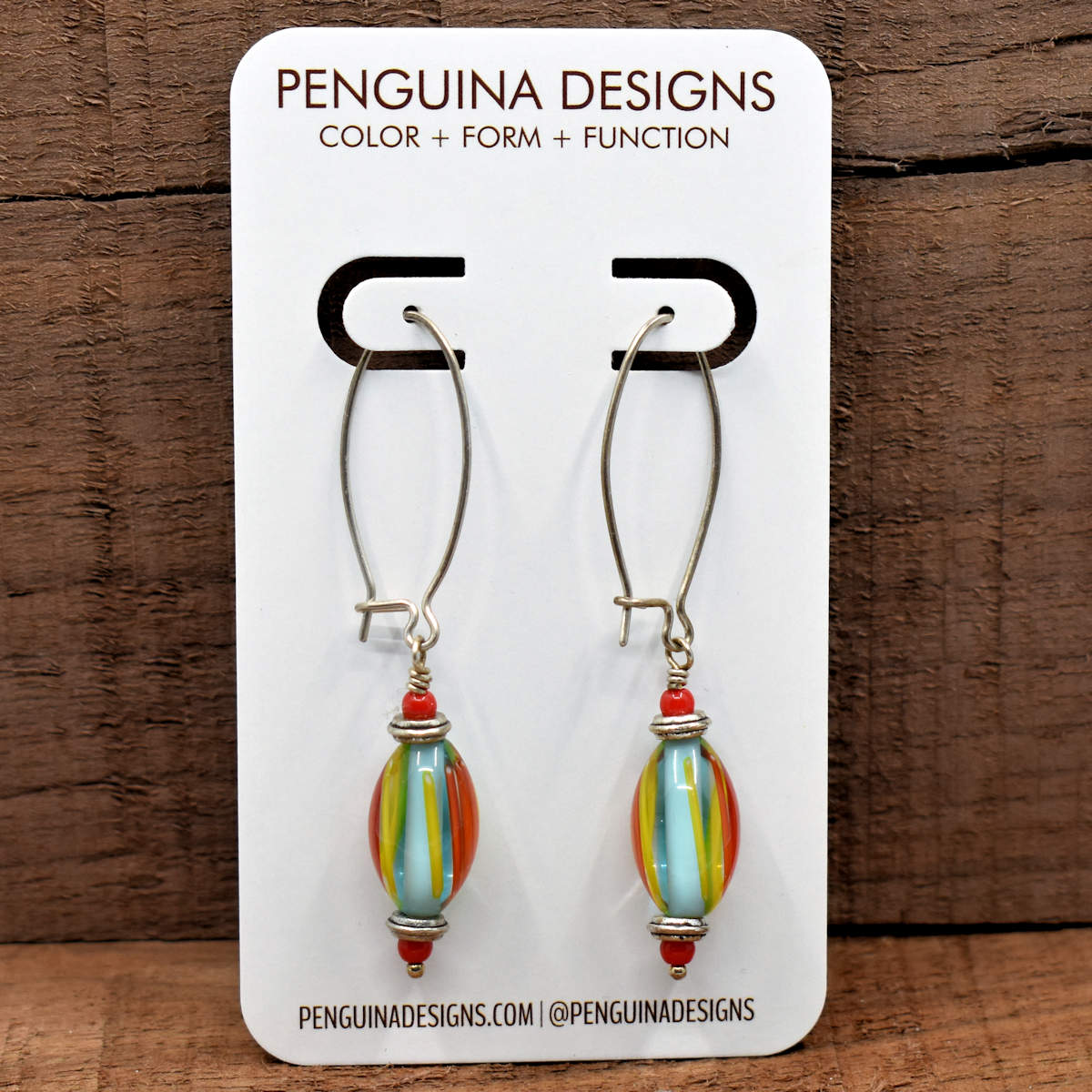 A pair of earrings on a white card rest against a wood background. The earrings have long silver oval wires that latch and the oval drops at the bottom are a light blue with orange and red stripes.