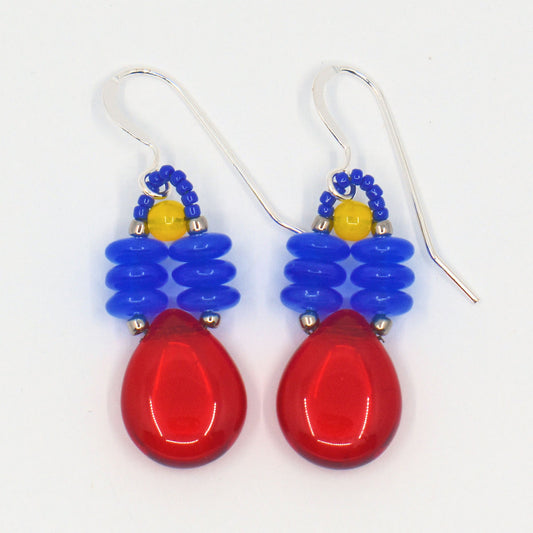 A pair of brightly colored earrings laying on a white background. The earrings have a big clear red teardrop suspended from two parallel stacks of blue discs.