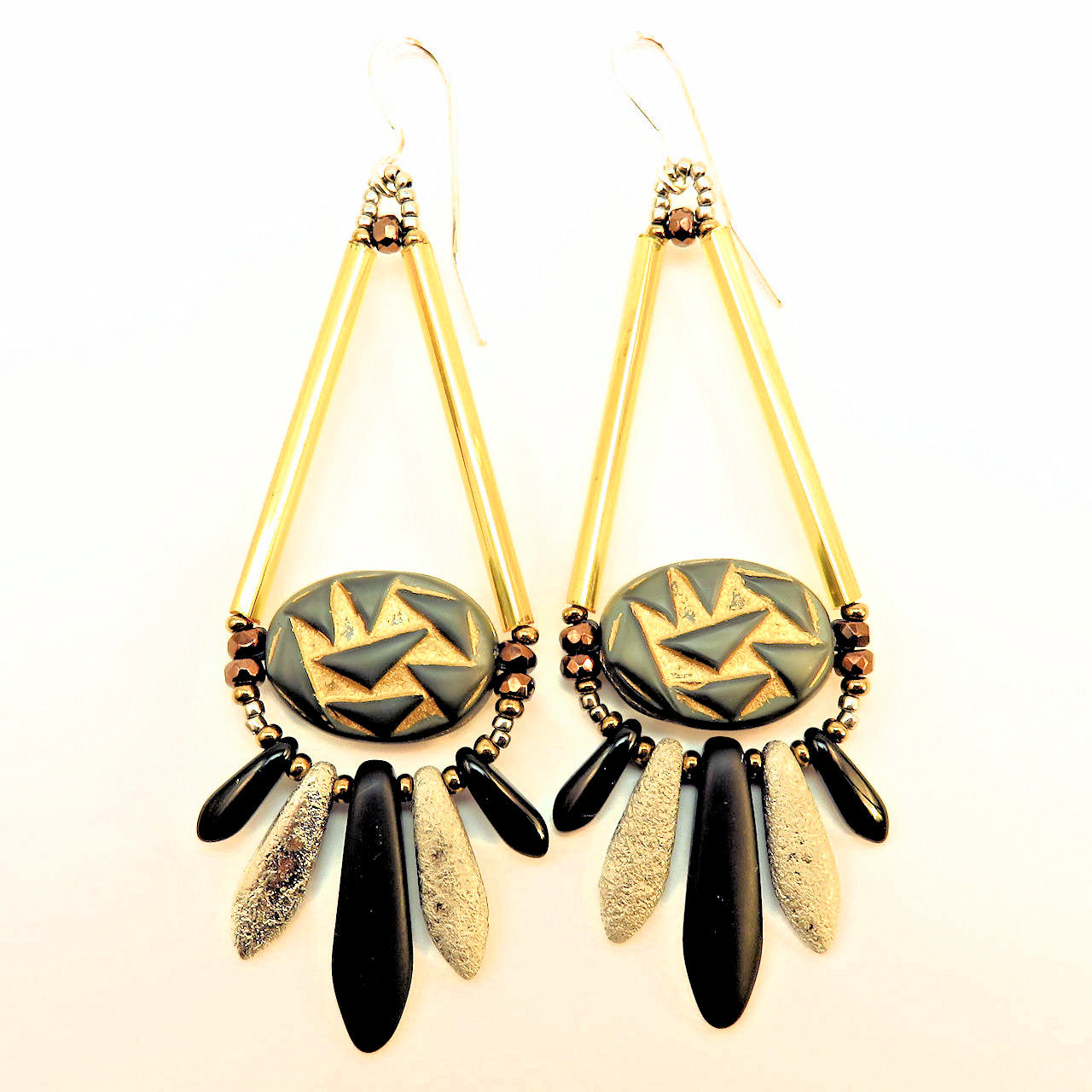 Teardrop shaped earrings in gold, silver, bronze and black lay on a white background. The earrings are an open teardrop shape, with the wide part filled by silvery gray ovals with an abstract triangle design that is antiqued in gold. Below the teardrop are five dagger beads, small black on the outside, a textured silver inside, and a large matte black one in the center. 