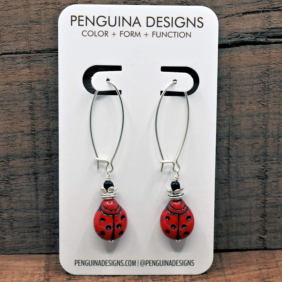 A pair of earrings on a white card rest against a wood background. The earrings have long silver oval wires that latch and have red glass lady bugs at the bottom.
