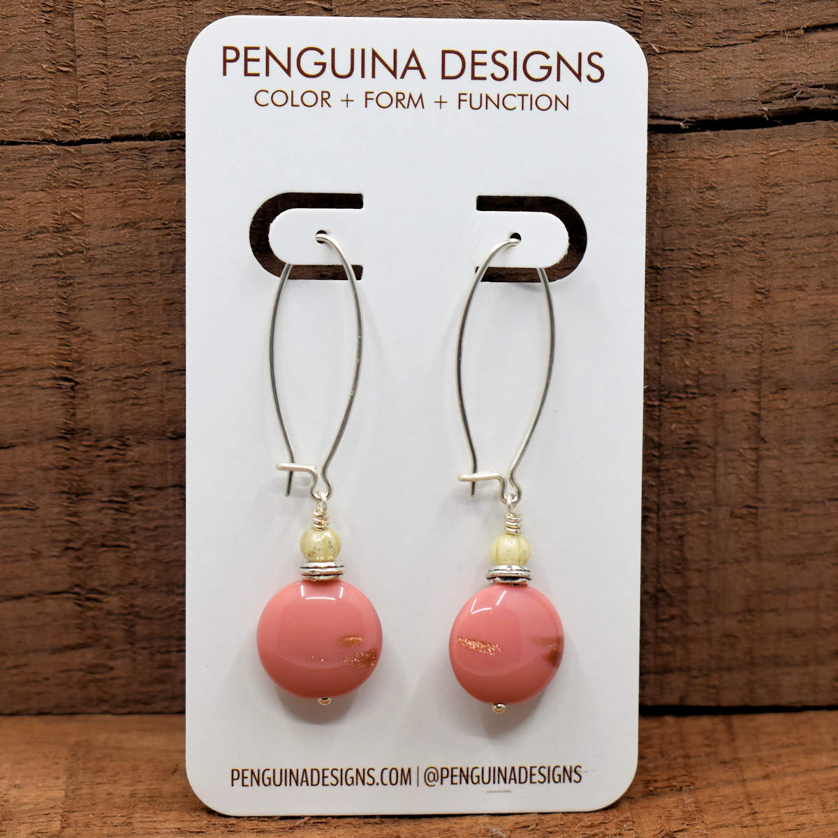 A pair of earrings on a white card rest against a wood background. The earrings have long silver oval wires that latch and pink discs with little bits of coppery glitter at the bottom.