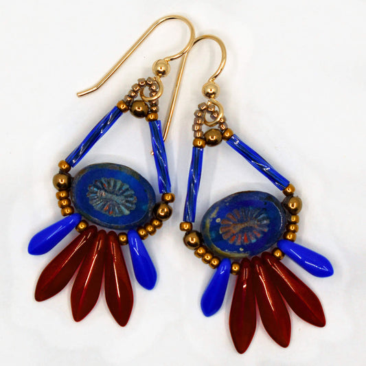 Dark blue and dark red earrings with gold wires lay on a white background. They are teardrop shaped, with blue tubes forming the upper straight sides and a blue oval bead with an embossed starburst pattern in the middle. At the bottom of the teardrop shape is a fan of dagger beads, small opaque blue ones on the outside and three dark red ones in the middle.