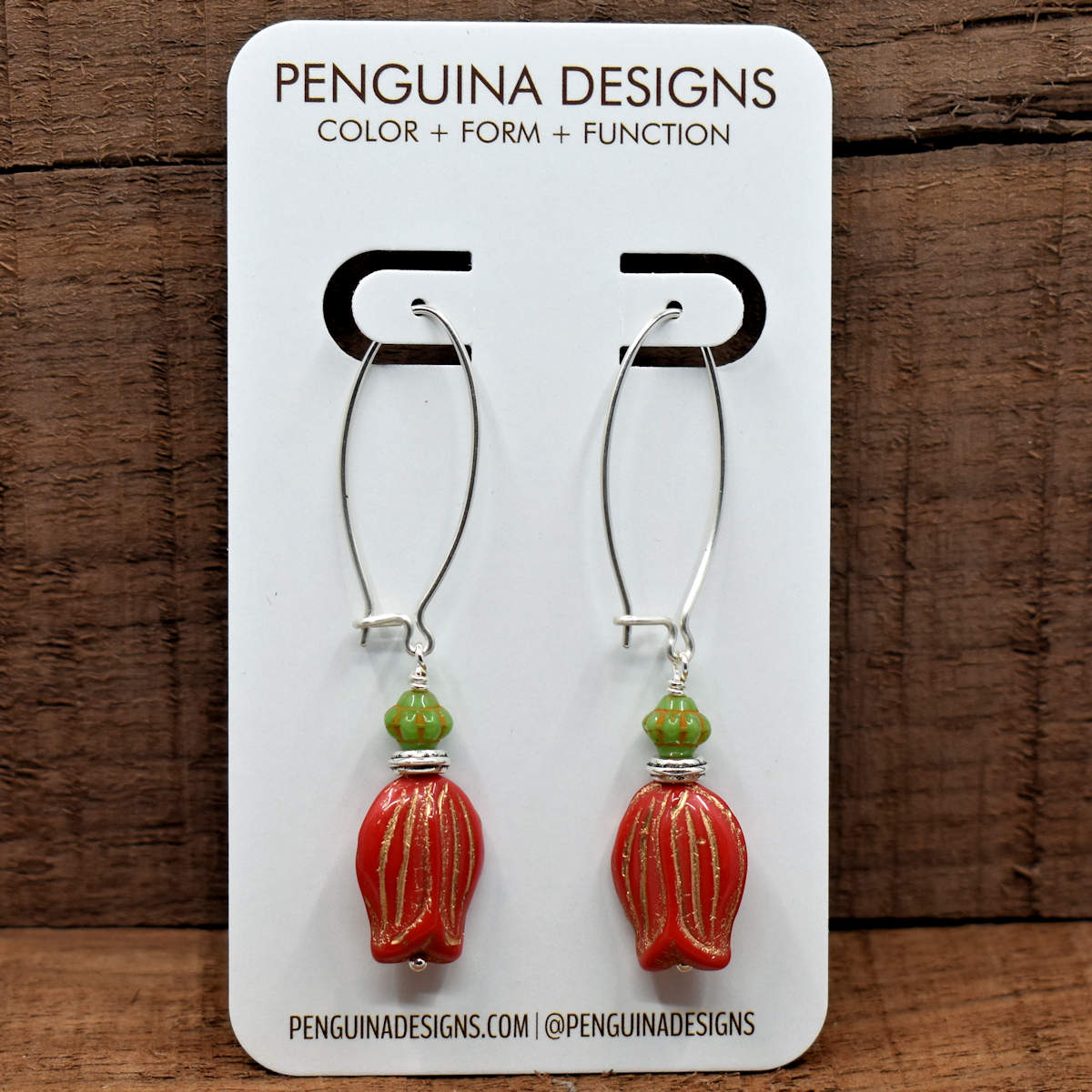 A pair of earrings on a white card rest against a wood background. The earrings have long silver oval wires that latch and at the bottom there are red upside down tulip beads with a green bead on top of them.