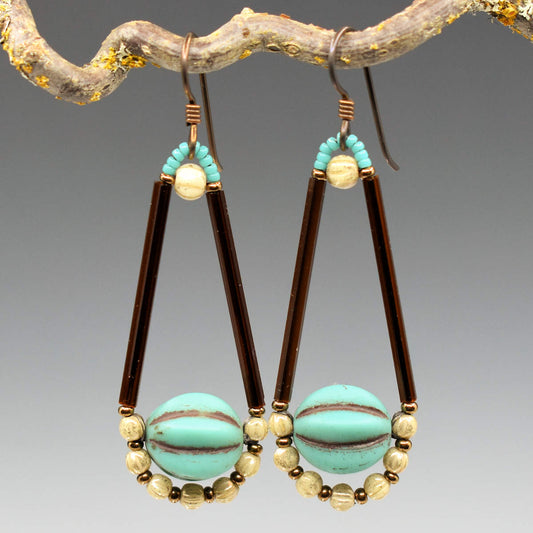 Long teardrop shaped earrings with long brown tubes and turquoise balls at their base hang from a twig. The earrings have an arc of small cream beads around the bottom of the large turquoise bead and dark metal ear wires.