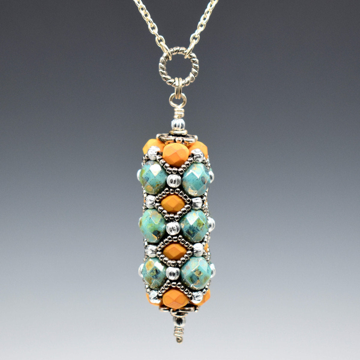 A pendant with orange and turquoise hangs from a rope textured circle and silver colored chain. The pendant is a column of bronzed turquoise faceted beads alternating with soft orange beads, all of which are overlaid with a silver seed bead netting. 