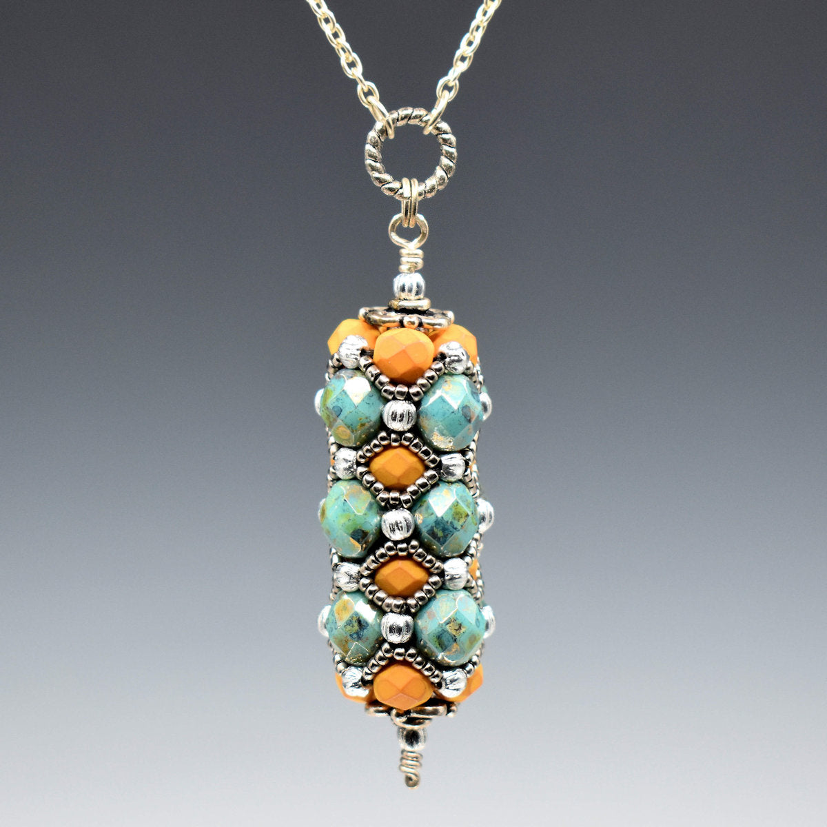 A pendant with orange and turquoise hangs from a rope textured circle and silver colored chain. The pendant is a column of bronzed turquoise faceted beads alternating with soft orange beads, all of which are overlaid with a silver seed bead netting. 