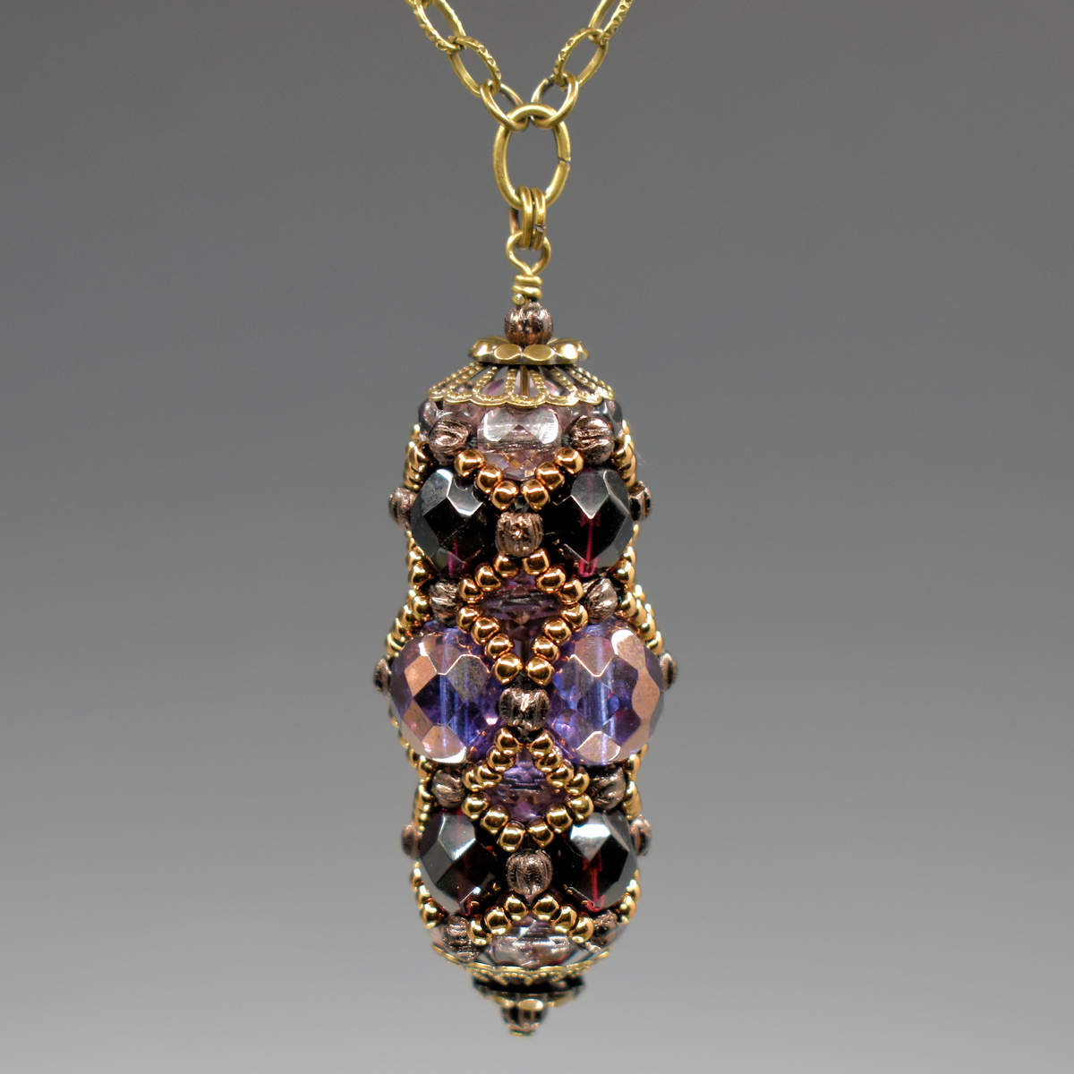 A purple and gold beaded pedant on gold colored chain. The pendant has larger light purple faceted beads in the center row and darker, warmer purple beads in the rows on either side. The whole pendant is overlaid with a netting of gold seed beads and capped with filigree style gold colored metal.