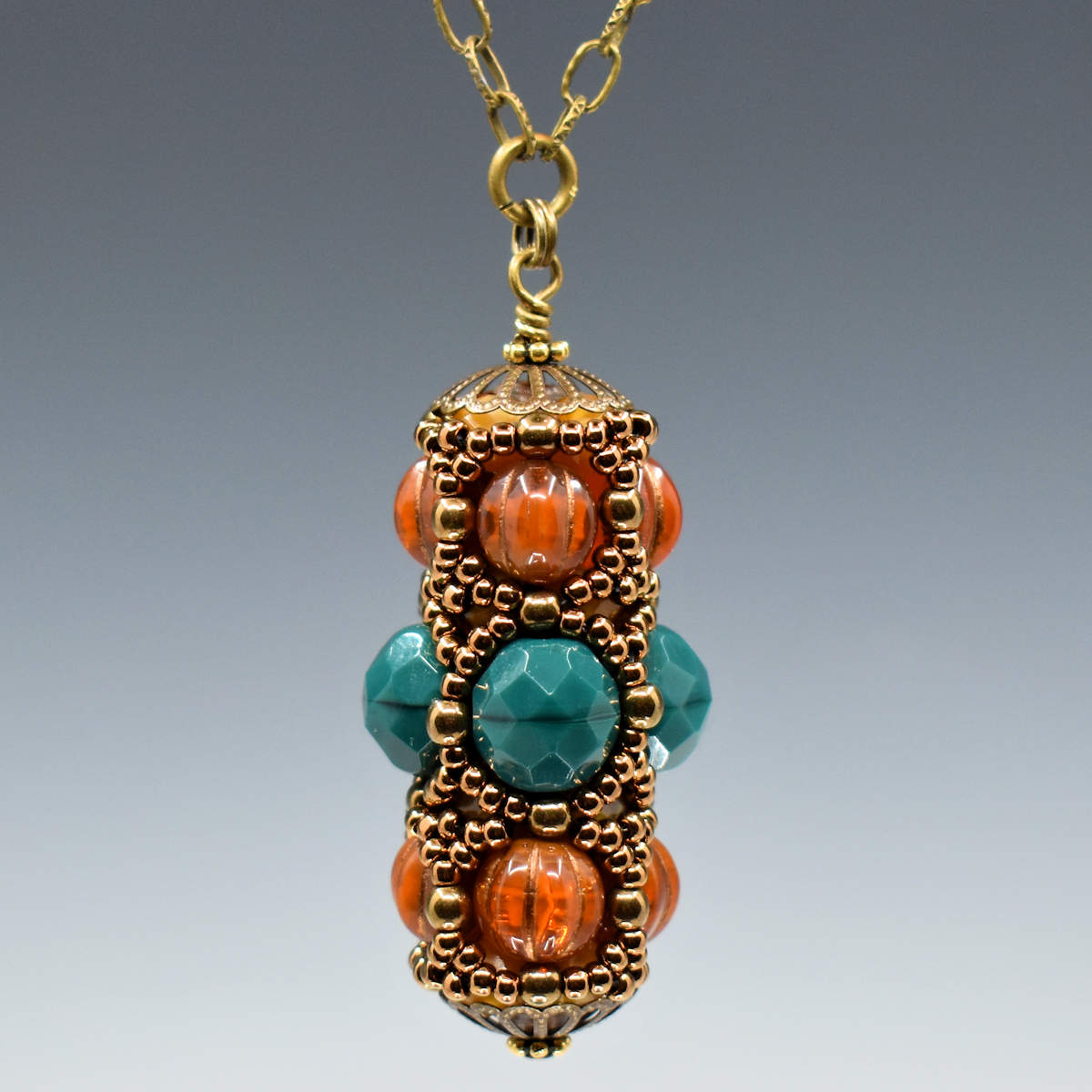 A forest green and rust brown pendant hanging from gold colored chain. The pendant is wider in the middle, with green beads that have an internal mold stripe at their centers. On the upper and lower rows are ridged semi-transparent rust rounds. The whole pendant is overlaid with a netting of gold seed beads and capped with golden filigree style findings.