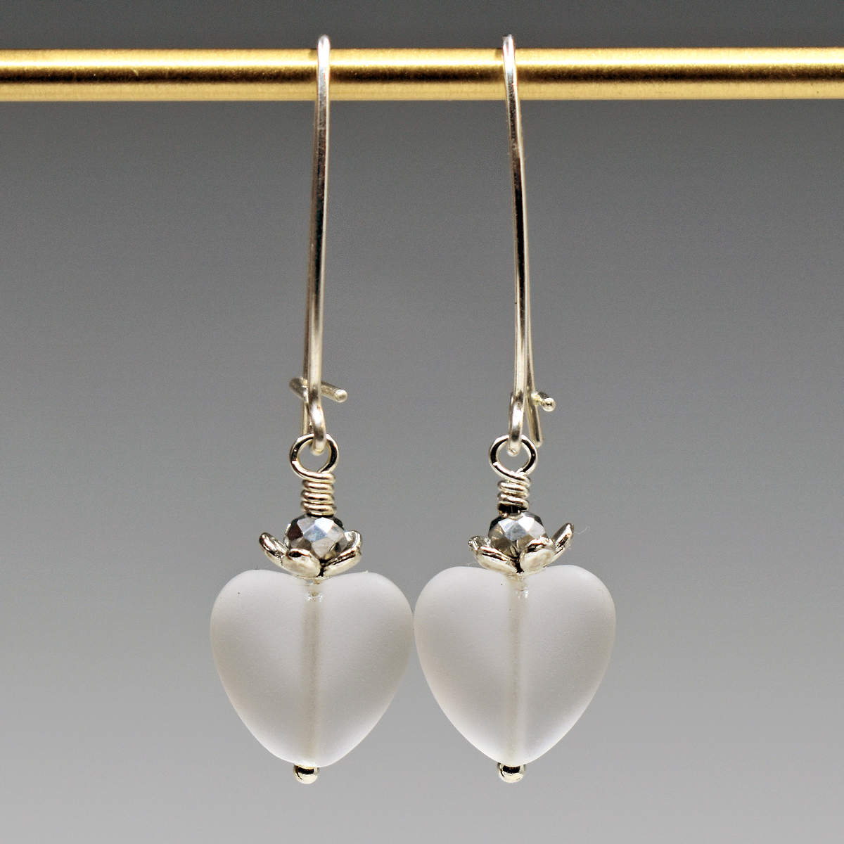 A pair of earrings hang from a gold bar, against a gray background. The earrings have long silver oval wires that latch and small frosted clear glass hearts at the bottom.