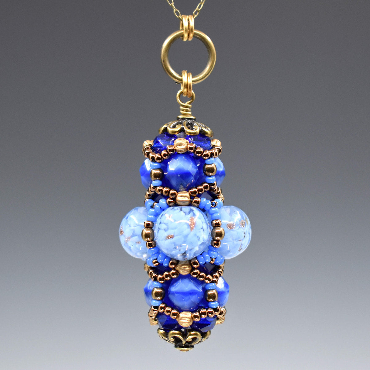 A blue and gold pendant hanging from a gold colored chain against a gray background. The middle of the pendant has clear beads filled with flecks of light blue and some gold glitter accents. The top and bottom layers are made from smaller medium blue beads. The whole pendant is overlaid with gold and blue seed beads. 