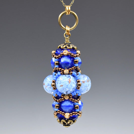 A blue and gold pendant hanging from a gold colored chain against a gray background. The middle of the pendant has clear beads filled with flecks of light blue and some gold glitter accents. The top and bottom layers are made from smaller medium blue beads. The whole pendant is overlaid with gold and blue seed beads. 