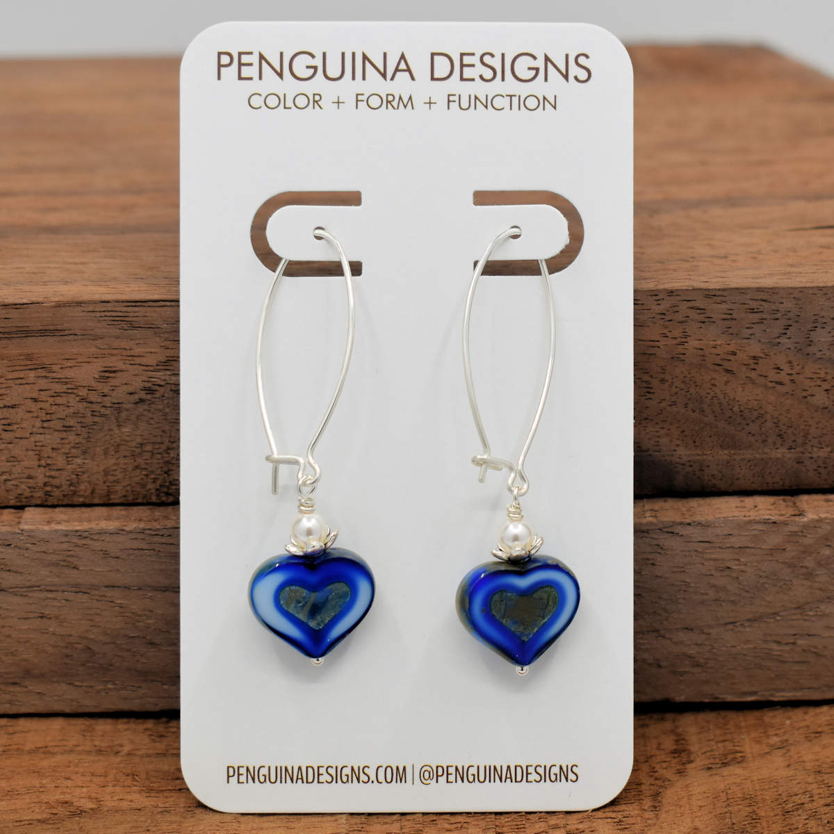 A pair of earrings on a white card rest against a wood background. The earrings have long silver oval wires that latch and blue hearts at the bottom with a white pearl bead. The blue hearts are a variegated dark and light blue and have a darker stonelike texture heart  pressed into the center.