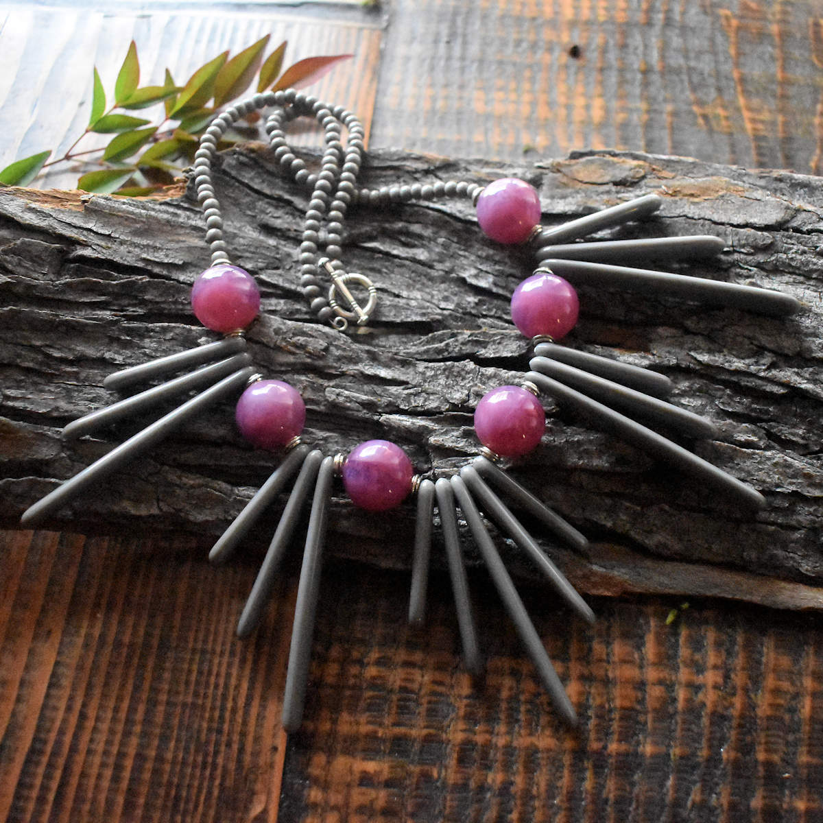 A necklace with gray fringe beads and purple round beads is draped across a wood backdrop. The necklace has five sections of gray spine beads, with the outer two sets grouped in inwardly ascending groups of three and the center section of five spines increasing from shortest at the outer sides to longest in the very center. Six big warm purple beads alternate with the gray sections. The necklace has a silver toggle clasp and smaller gray beads that form the back part of the necklace.