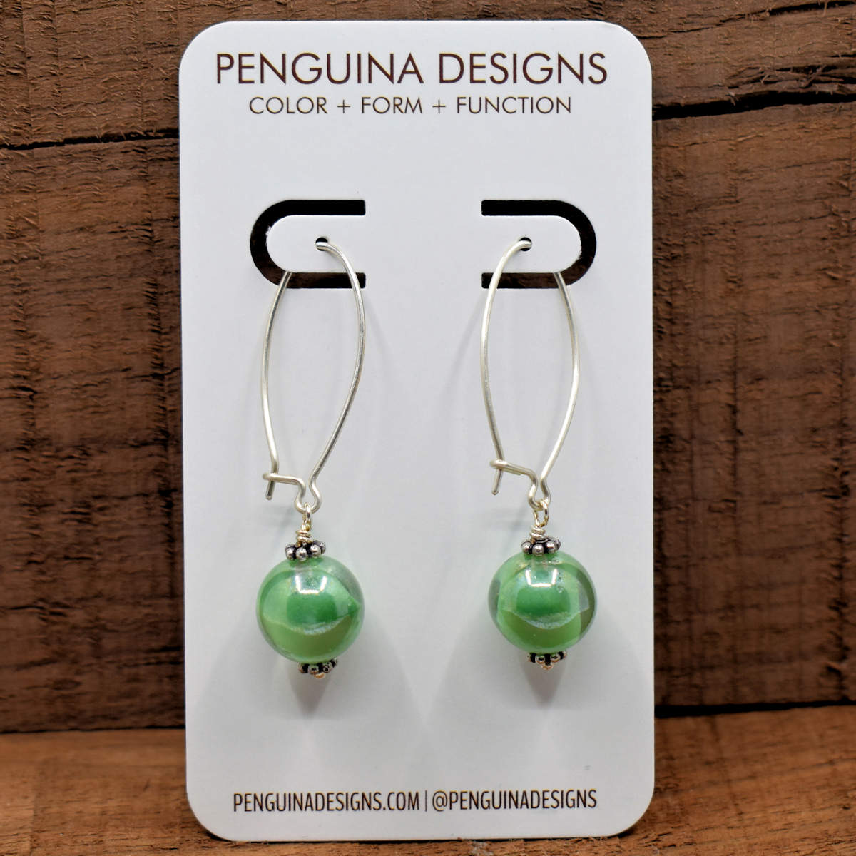 A pair of earrings on a white card rest against a wood background. The earrings have long silver oval wires that latch and glass balls with swirled green and clear glass at the bottom.