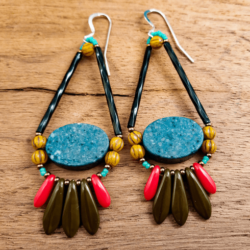 Long, colorful teardrop shaped earrings with silver ear wires lay on a wood background. The earrings' teardrop shape has a long twisted black tube for each straight side with two mustard colored round beads below it. There is a speckled dark turquoise oval in the wide lower part of the teardrop and at the bottom is a swag of three dark greenish brown dagger beads sandwiched between smaller red daggers. 