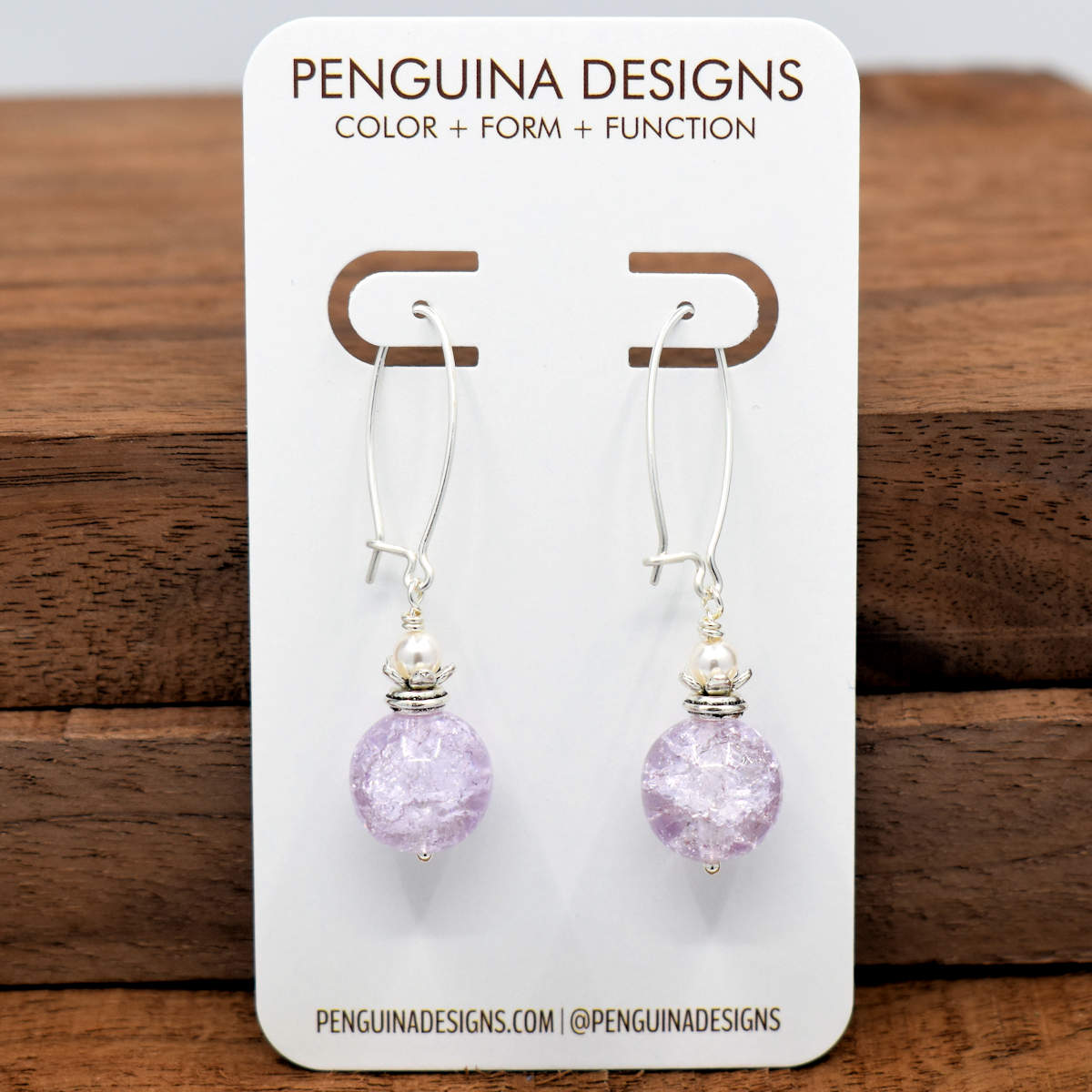 A pair of earrings on a white card rest against a wood background. The earrings have long silver oval wires that latch and pale lilac crackle glass rounds with a flower cup on top holding a white pearl bead. 