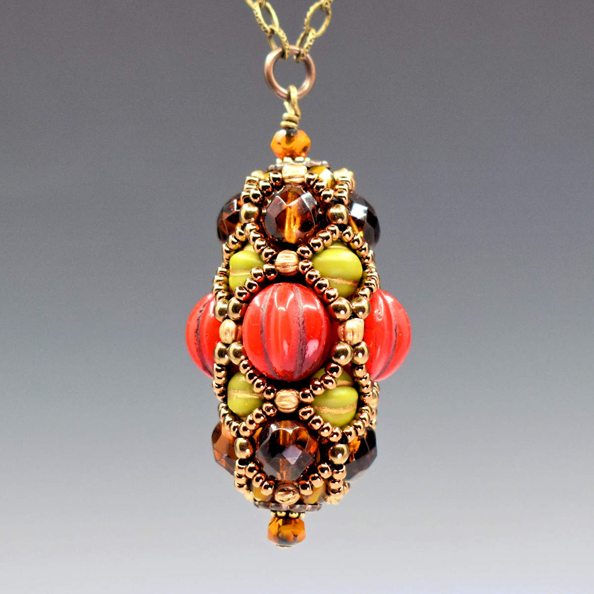 A beaded pendant that is egg shaped hangs from a gold colored chain, against a gray background. The widest part of the pendant is ridged warm red beads, and then ridged avocado beads are nestled between the red beads and an outer layer of varigated transparent amber beads. A netted layer of gold seed beads is overlaid, outlining the red, green, and brown beads. 