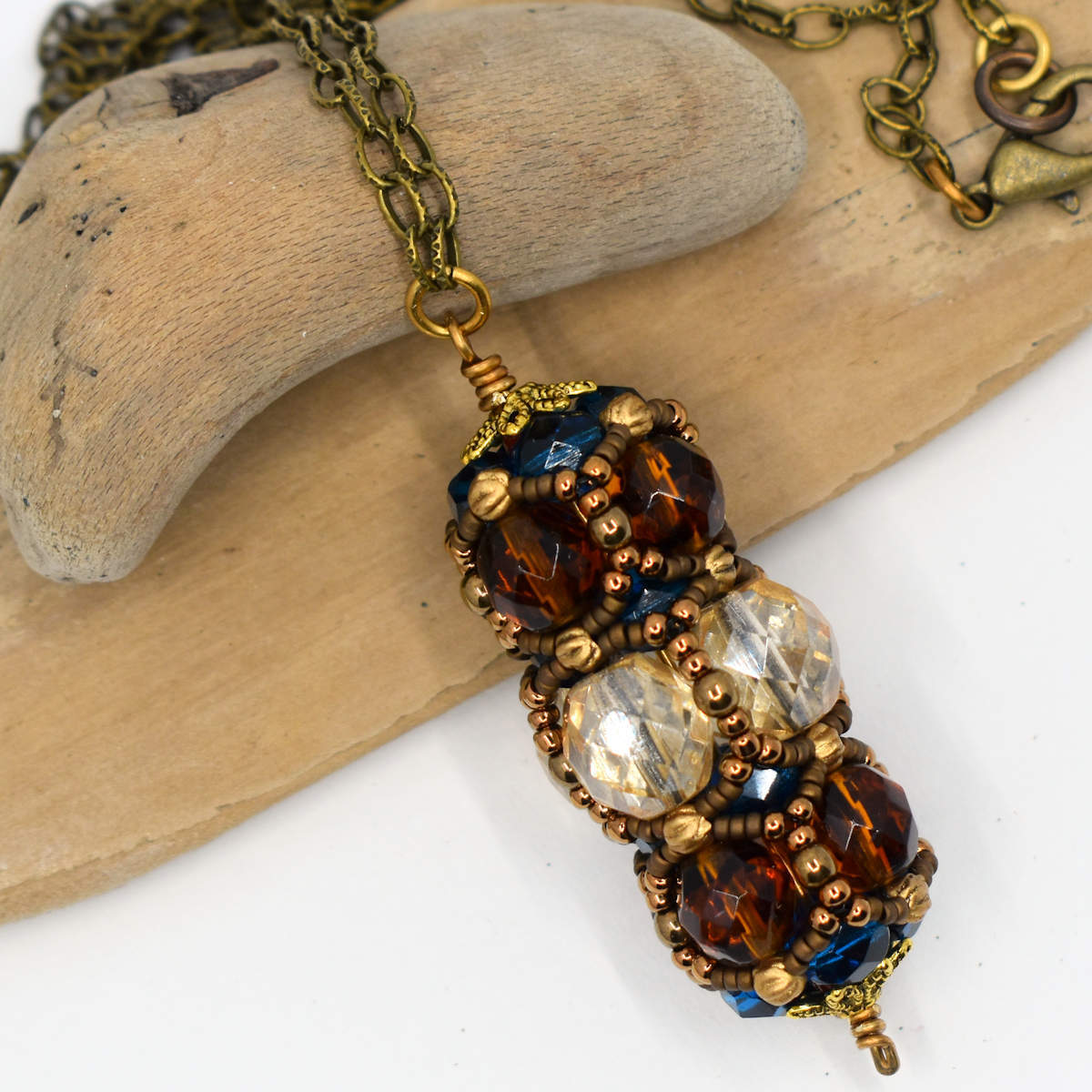 A columnar brown and clear beaded pendant is draped against smooth driftwood. The pendant has a center row of slightly yellowy clear faceted beads and the two outer layers are variegated dark amber.  Through the overlay of matte and shiny gold bead netting, dark capri blue beads are just visible at the top and bottom of the pendant. 
