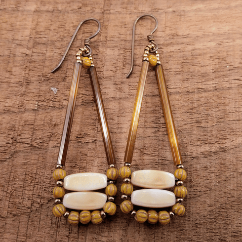 Elongated triangle earrings in brown and cream lay on a wood background. The triangle shape open at the top, with long brown tube beads outlining it. The bottom is formed from two horizontal cream rectangles that are outlined by small mustard colored ridged rounds alternating with gold seed beads. 
