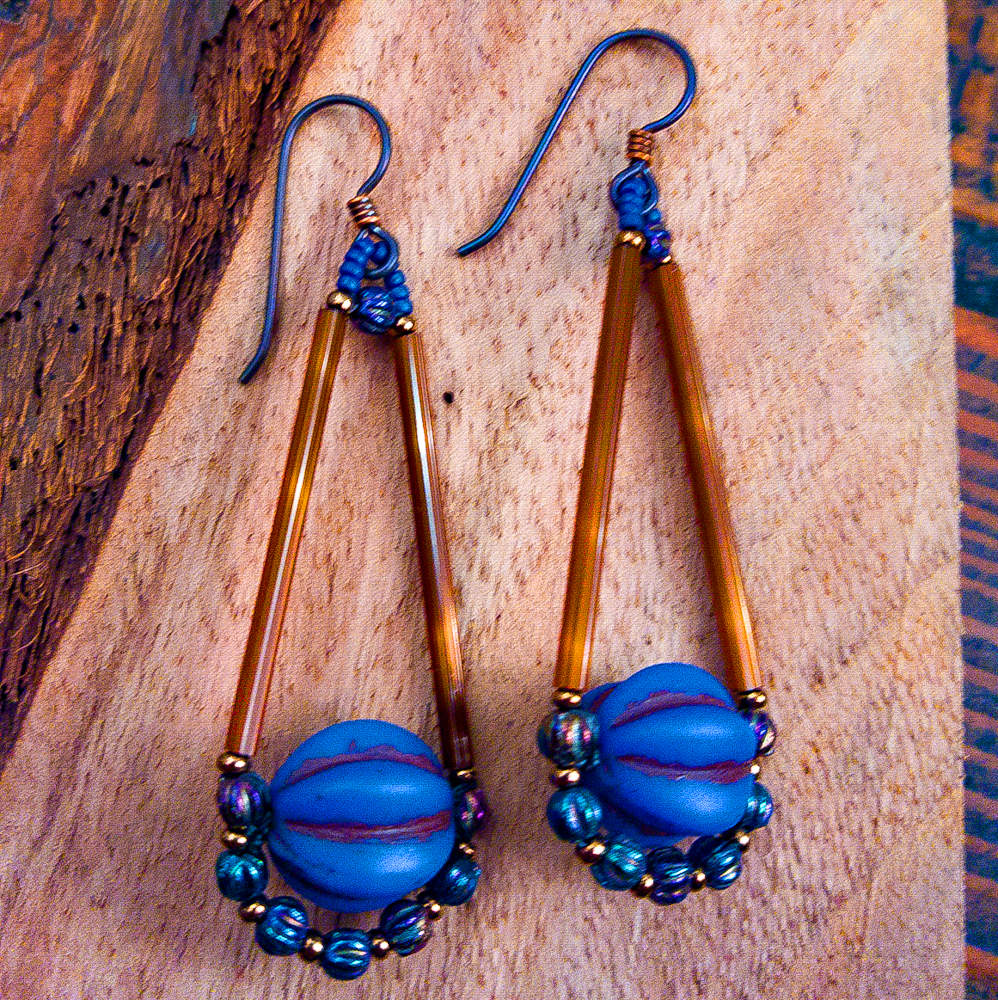A pair of brown and cobalt blue teardrop shaped earrings lay on wood. The teardrop shape of the earrings is formed with two long brown beads on the upper, straight sides, and at the bottom is a matte blue bead with deep horizontal ridges. The bottom of the teardrop is outlined by small iridescent blue rounds alternating with gold seed beads. The ear wires are dark metal. 