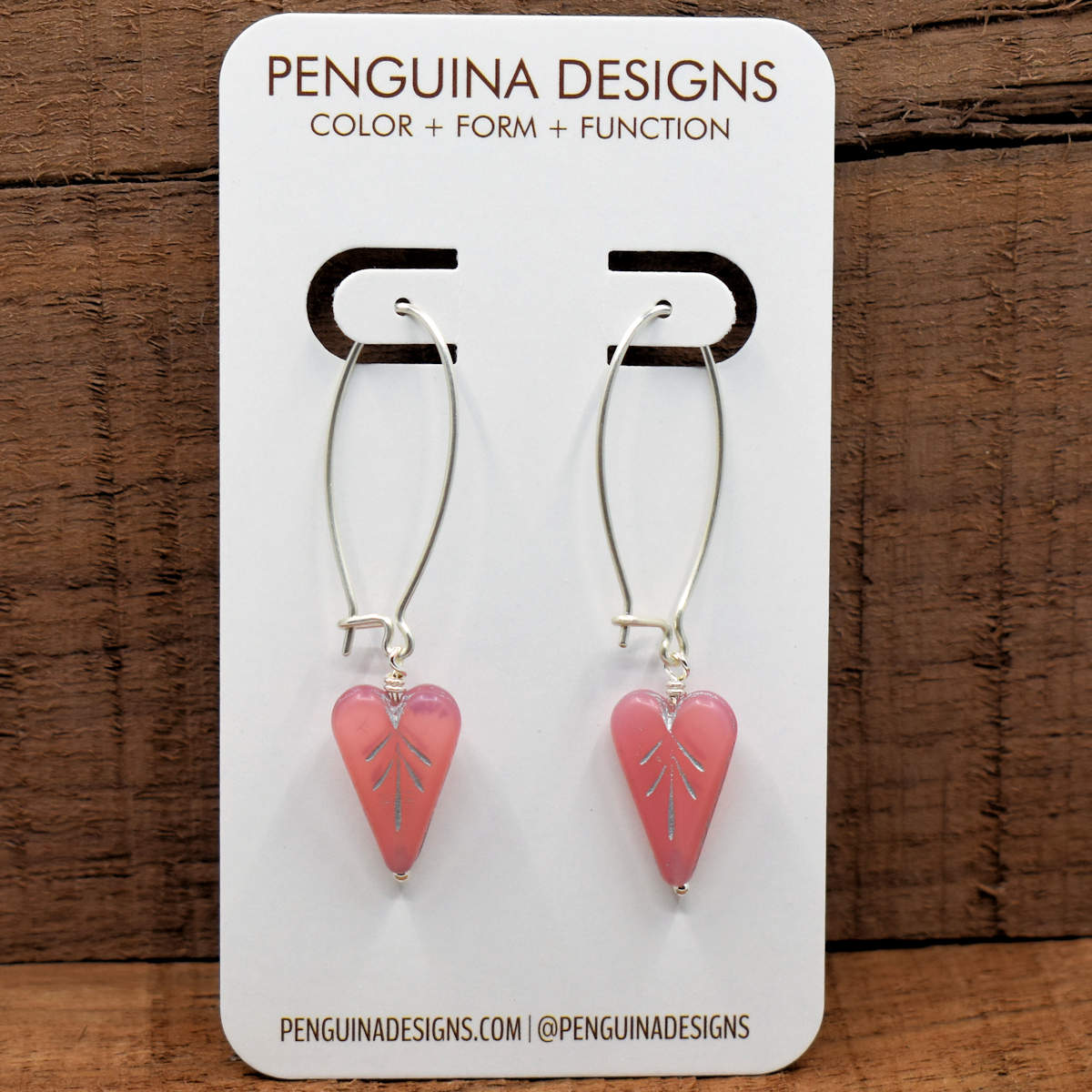 A pair of earrings on a white card rest against a wood background. The earrings have long silver oval wires that latch and slightly translucent pink elongated hearts at the bottom.