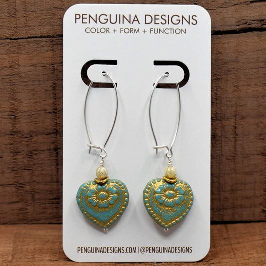 A pair of earrings on a white card rest against a wood background. The earrings have long silver oval wires that latch and the bottom dangles are turquoise hearts embossed with flower designs.