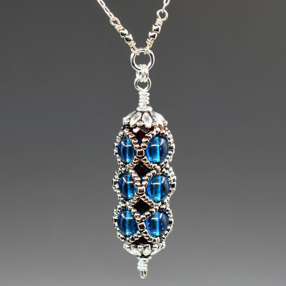 A capri blue and silver pendant against a gray background. The pendant is a very columnar shape made from smooth round capri blue beads. There is an overlaid netting of silver seed beads and the pendant is capped with a silver flower bead cap.