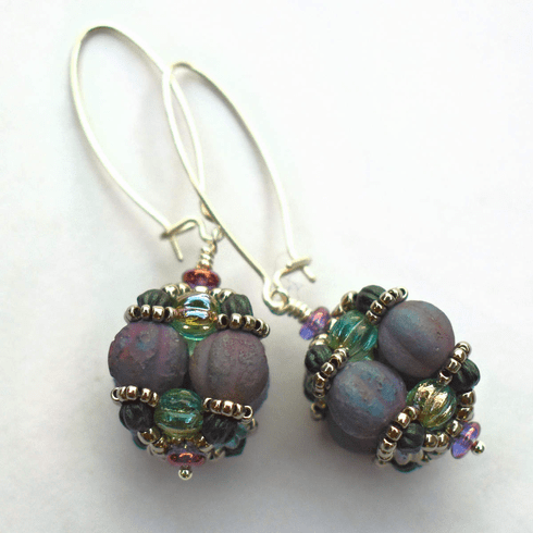 A pair of earrings laying against a white background. The earrings have long silver oval wires that latch and there is a beaded ball dangling from the bottom of each earring. The beaded beads are s made from rough textured lilac beads surrounded by layers of silver and sea green beads. 