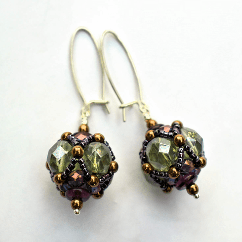 A pair of earrings laying against a white background. The earrings have long silver oval wires that latch and there is a beaded ball dangling from the bottom of each earring. The beaded beads are made from pale transparent green beads that have a layer of netted putple and bronze beads wrapped around them.