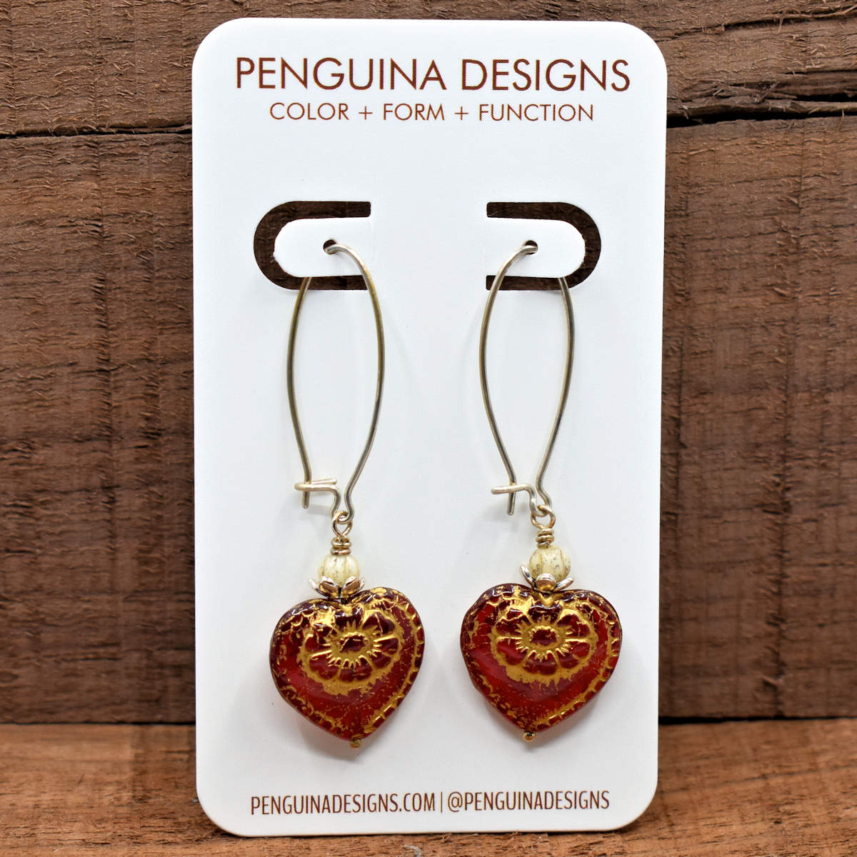 A pair of earrings on a white card rest against a wood background. The earrings have long silver oval wires that latch and red hearts at the bottom that are embossed with a gold flower design.