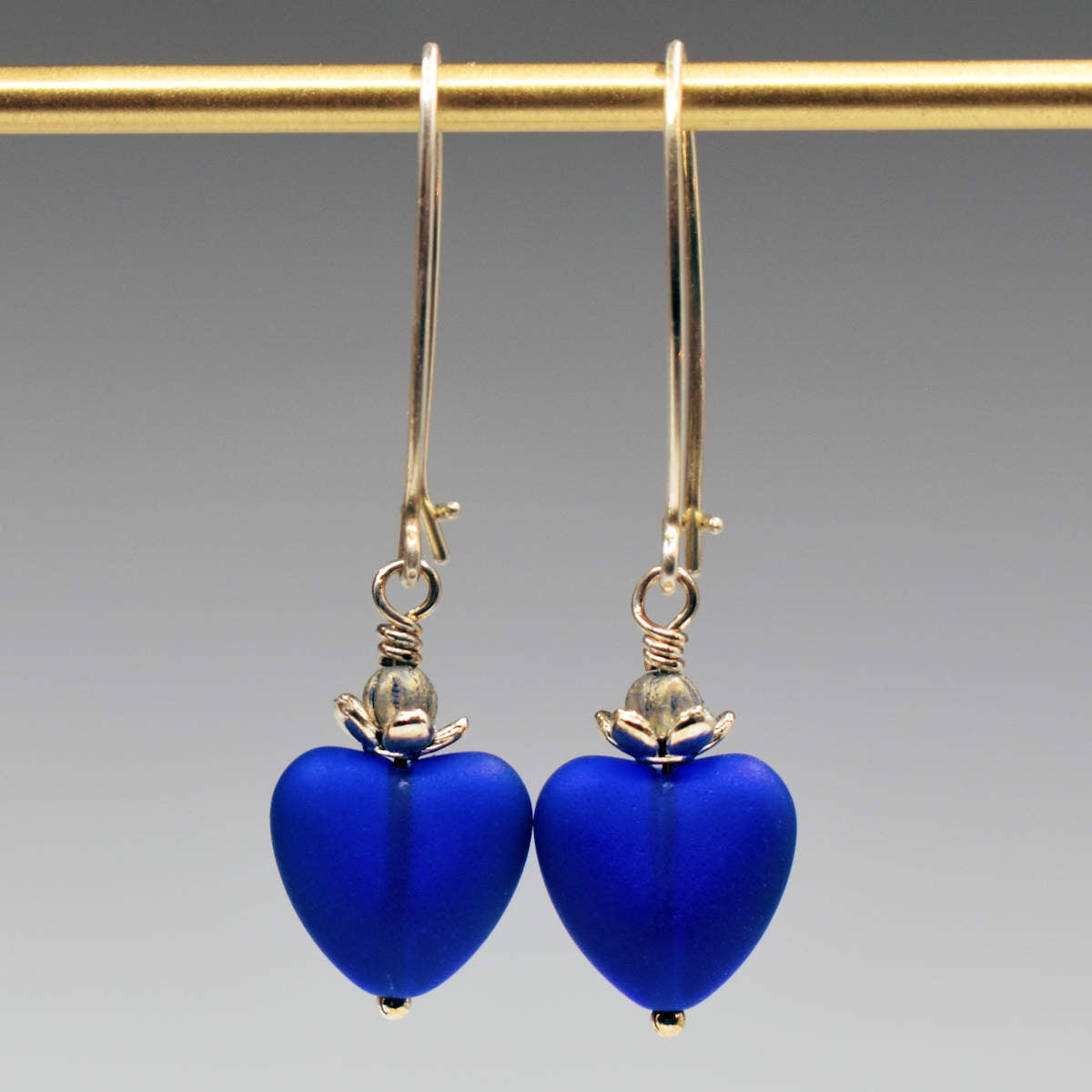 A pair of earrings hand from a gold bar, against a gray background. The earrings have long silver oval wires that latch and have drops that are made from small frosted cobalt glass hearts topped by a flower shaped cup that holds a silvery bead.