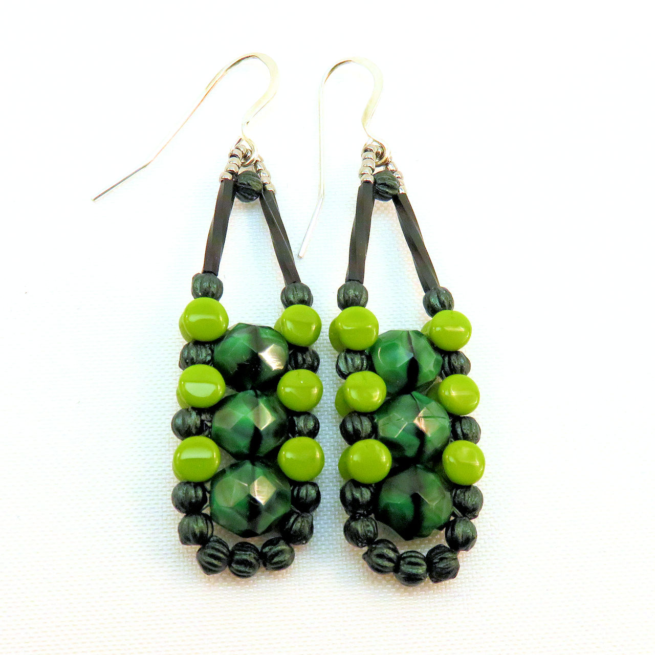 Long earrings in different shades of green with silver accents and ear wires on a white background. There are three swirled medium green beads stacked vertically in the center of these beads, like peas, surrounded by an outline of dark metallic green and avocado green beads.