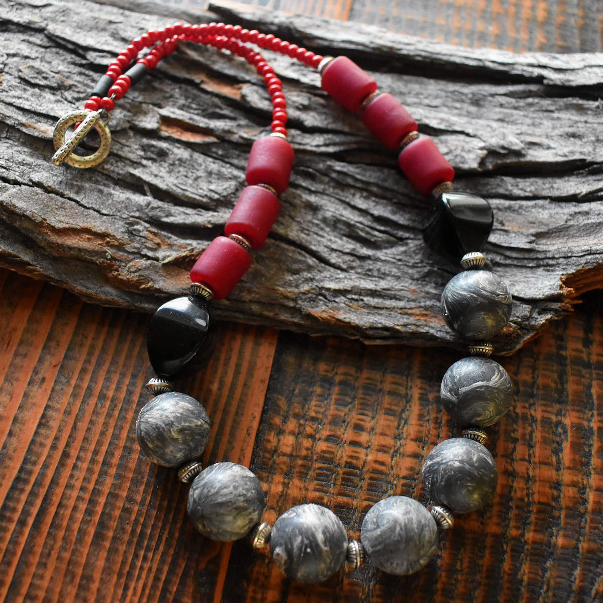 A gray, red, and black beaded necklace lays across a wood background. The necklace has seven gray and white swirled beads in the center, shiny black beads on either side, followed by three chunky matte red tube beads.  The back of the necklace is red seed beads with a silver toggle clasp.
