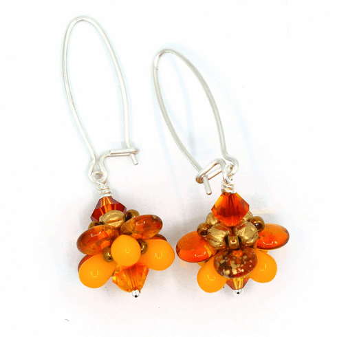 A pair of silver earrings with orange flower dangles on a white background. The earrings have long silver oval wires that latch and the dangling parts are orange beaded flowers with clear orange outer petals and bright light orange inner petals.