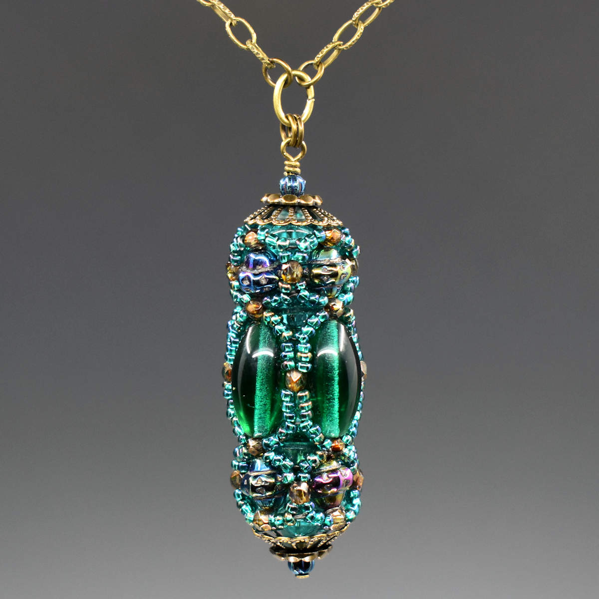 A turquoise and green beaded pendant hangs from a gold colored chain. The pendant has long oval beads that form the center row and textured iridescent beads that form a decorative top and bottom row. A netting of translucent peacock green seed beads is overlaid, outlining all of the larger beads. 