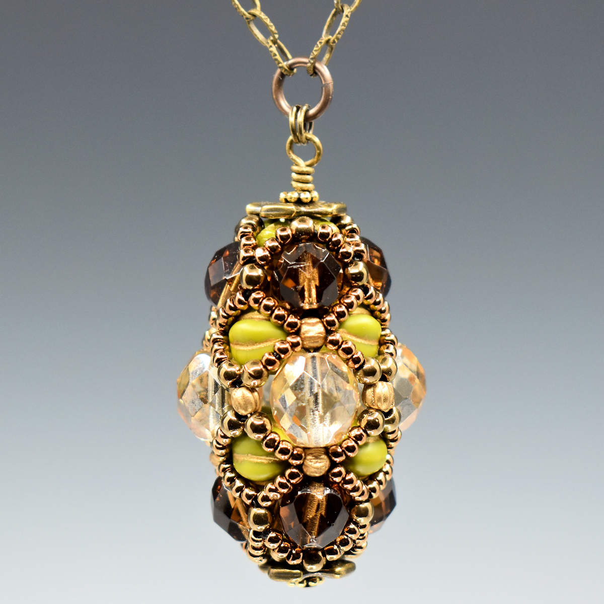 An egg shaped pendant hangs from a golden chain in front of a gray background. The pendant has a wide center section formed from transparent, faceted champagne colored beads, then a layer of matte avocado beads, and another of dark amber. A netting of gold seed beads is overlaid, outlining all of the larger beads. 